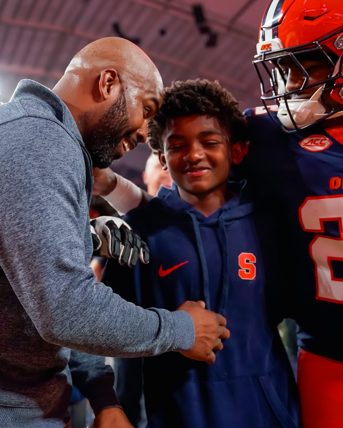 Two individuals wearing Syracuse University sports attire share a handshake. One person is in a coaching outfit while the other, a younger individual. They are smiling and seem to be enjoying a congratulatory moment.