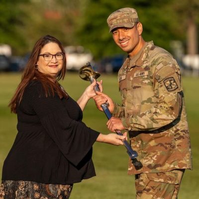 Two people participating in a ceremonial hammer handover on a grassy field, with one individual wearing U.S. Army camouflage uniform and the other in civilian attire.