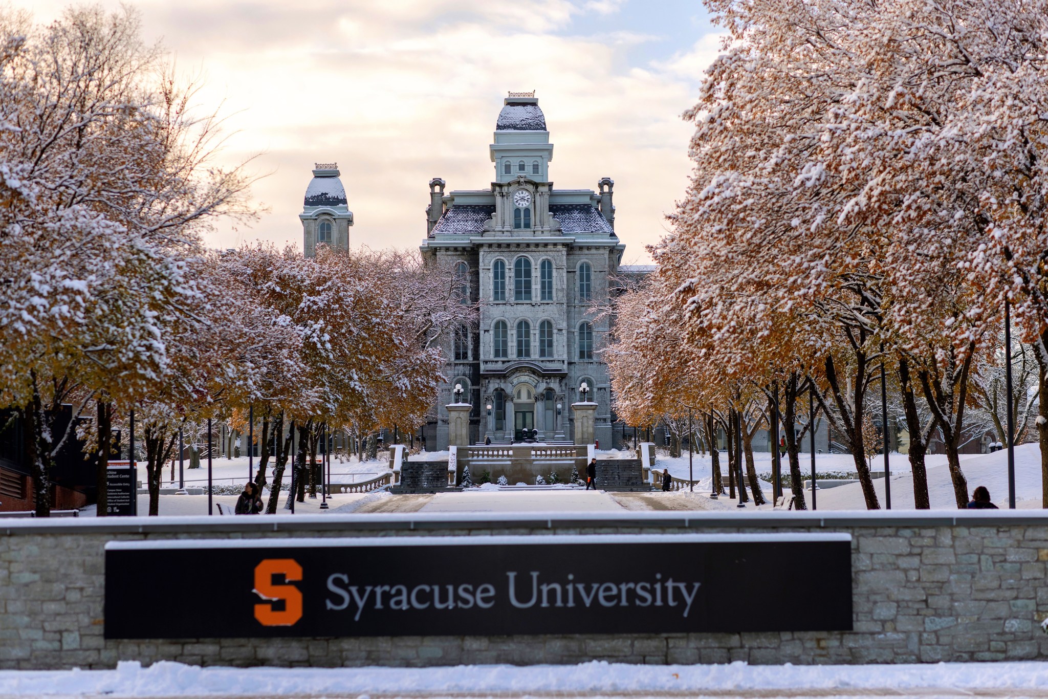 Snow-covered trees and grounds in front of the historic Hall of Languages building at Syracuse University, with the university’s name displayed on a sign in the foreground.