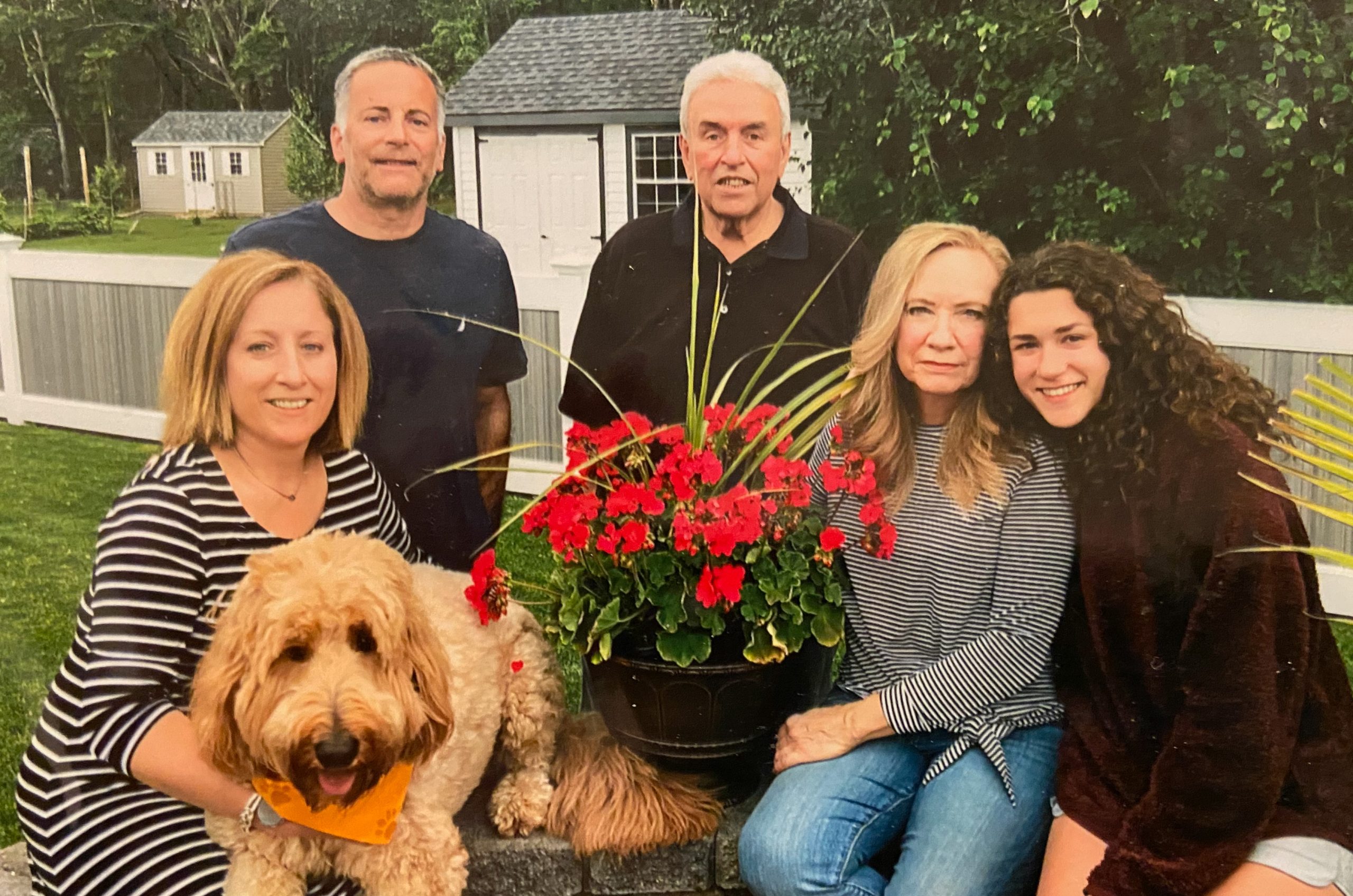 Five adults and a dog posing with a large pot of red flowers outdoors; no well-known entities visible.