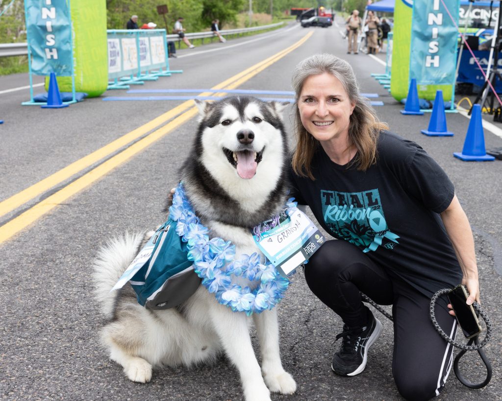 Person kneeling next to a large Alaskan Malamute at the finish line of a Teal Ribbon race, both looking happy. The dog wears a race bib and a lei.