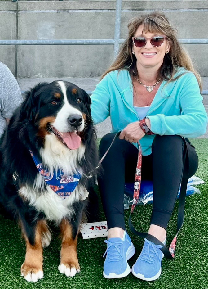 Person sitting on a step next to a large Bernese Mountain Dog, both outdoors, with the person smiling and wearing sunglasses.