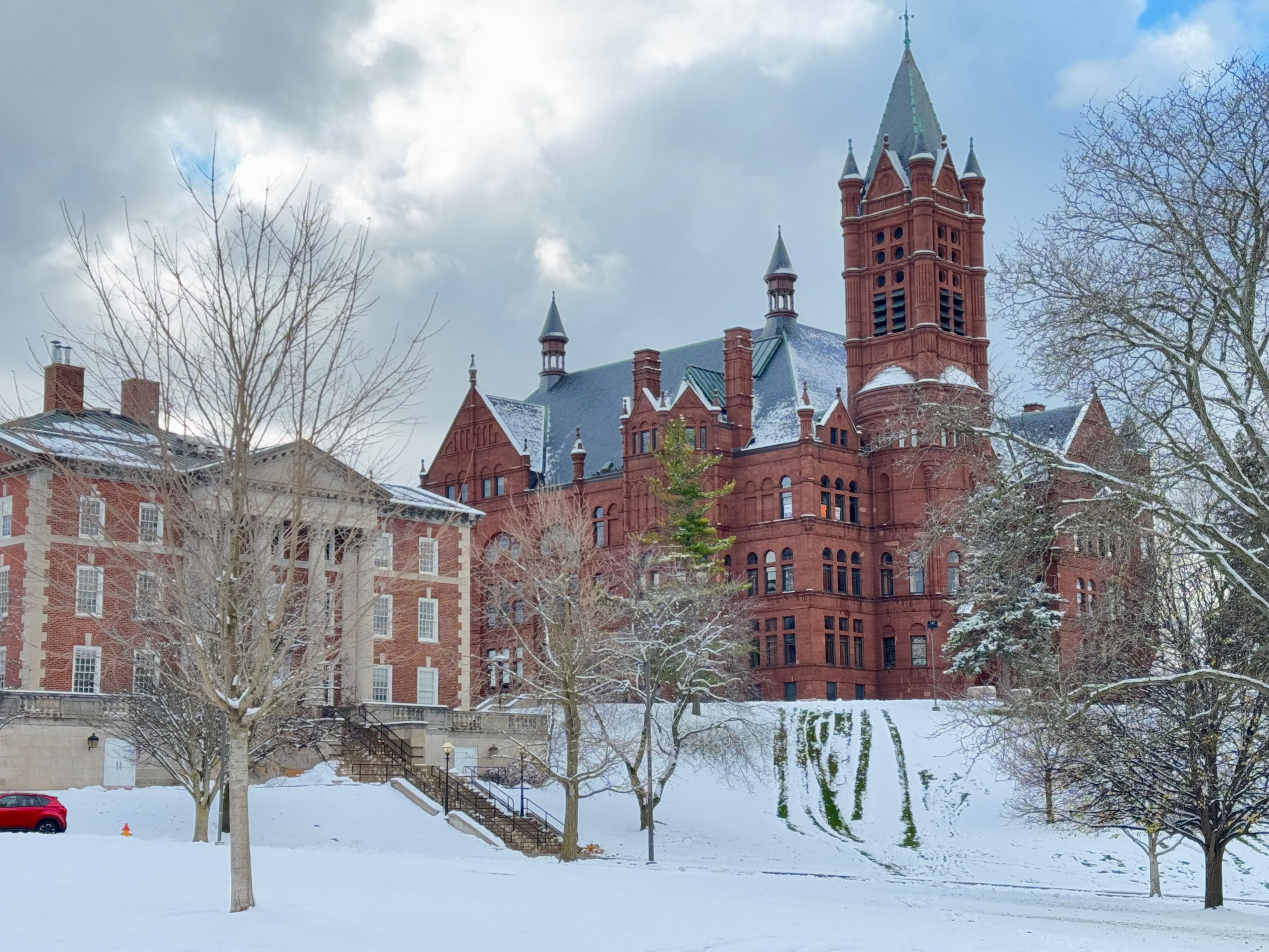 Snow covers the ground in front Crouse College, which features distinctive red brick architecture with pointed towers and historic detailing. Trees bare of leaves suggest a winter scene.