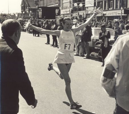 A woman wins the Boston Marathon, raising her hands up to her head as onlookers cheer her on.