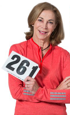 A woman smiles while holding up her number 261 Boston Marathon bib.