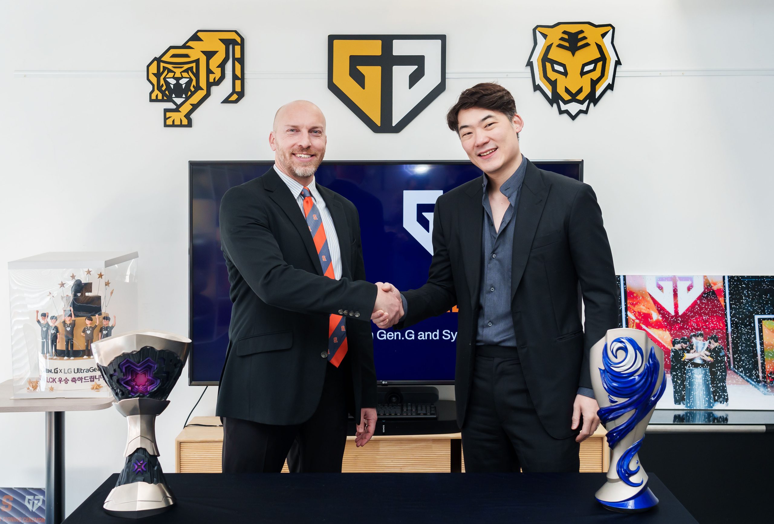 Two individuals shaking hands in front of a backdrop with Gen.G and G2 Esports logos, flanked by esports trophies.