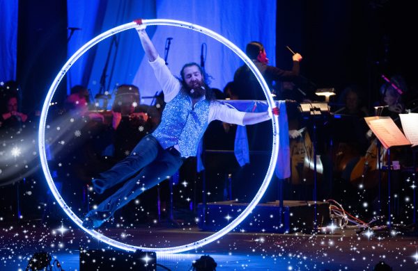 A performer in a sparkling blue outfit performs an aerial hoop act, suspended in front of a live orchestra on a dimly lit stage with twinkling lights in the background.