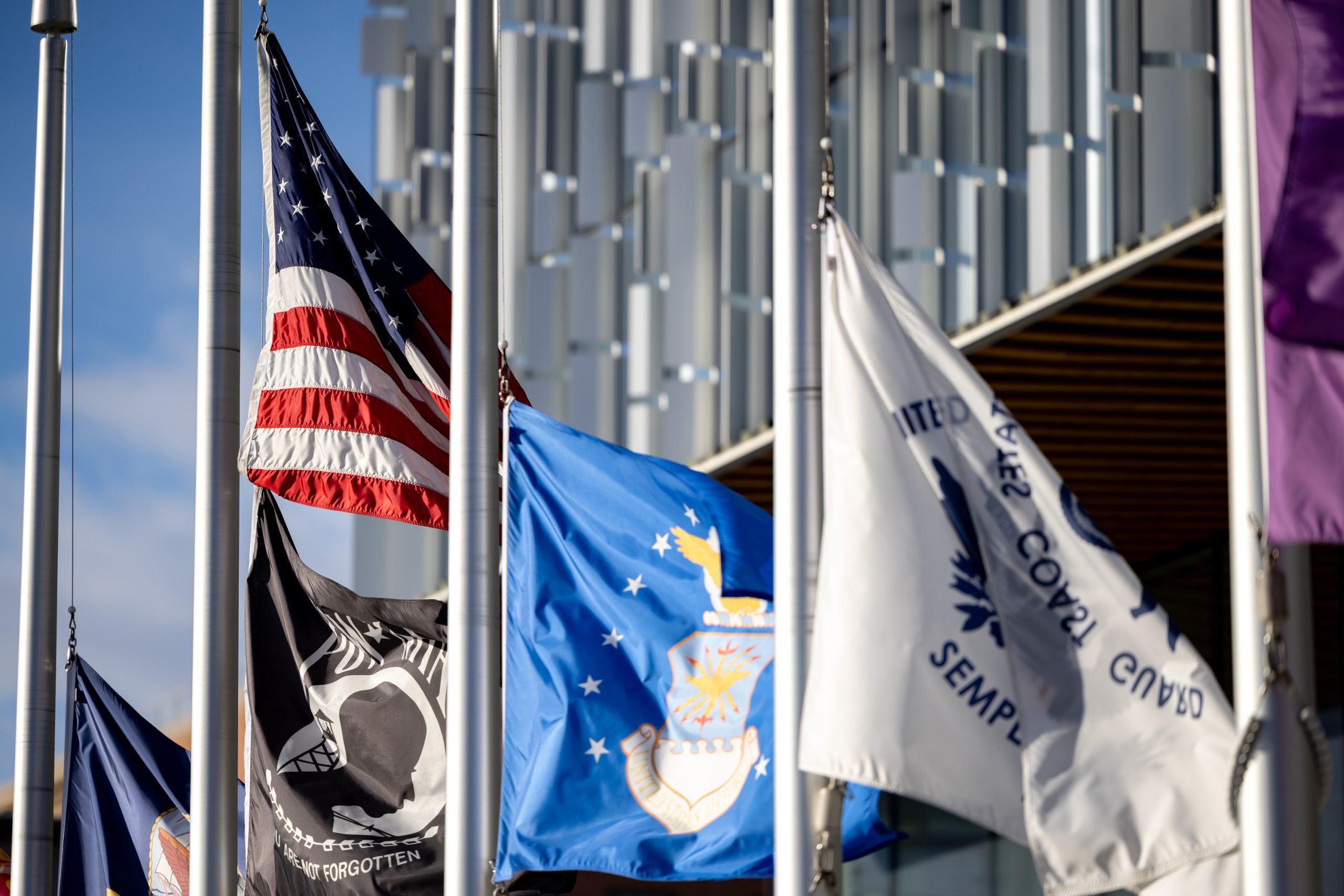 Flags, including the United States flag, the POW/MIA flag, and the United States Coast Guard flag, displayed in front of a modern building with vertical columns.