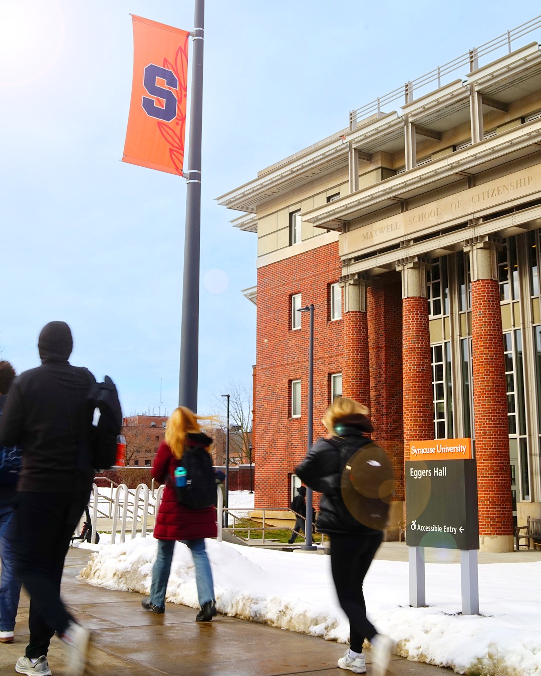 Pedestrians walking near the Maxwell School of Citizenship at Syracuse University, with an 'S' flag fluttering on a lamp post.