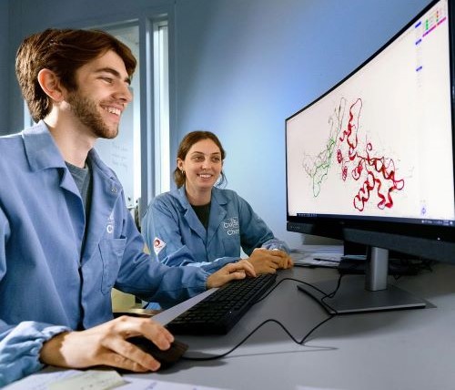 two students in blue lab coats look at a display of peptides on a computer screen