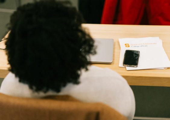 A person with curly hair sits at a desk with a laptop, smartphone, and Syracuse University College of Law folder.