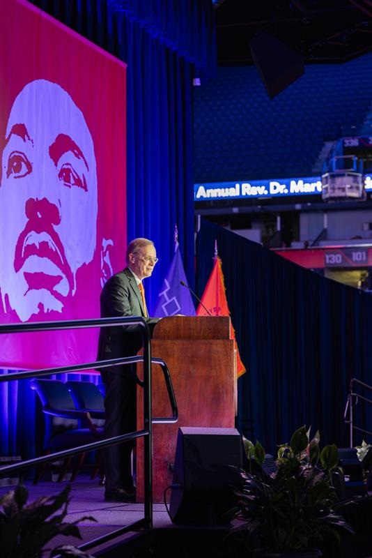 person speaking at a podium in JMA Wireless Dome in front of a banner with a graphic image 