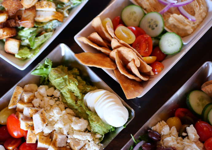 Various food items, including a salad with croutons, sliced eggs, veggies and hummus on plates.