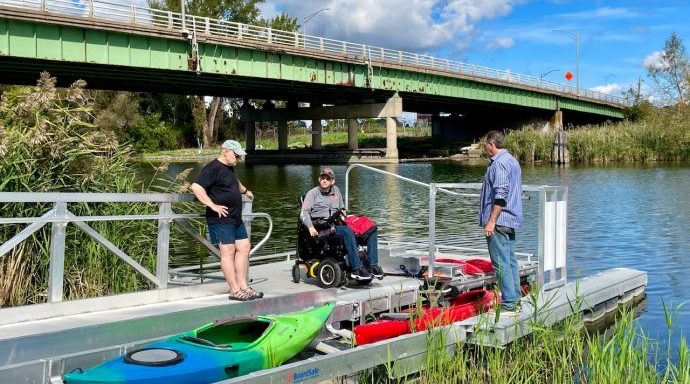 three people prepare to launch and board a kayak at an accessible ramp on the creek