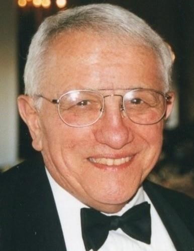 Portrait of an older individual with short gray hair and glasses, wearing a black tuxedo and bow tie, smiling against a warmly lit blurred background