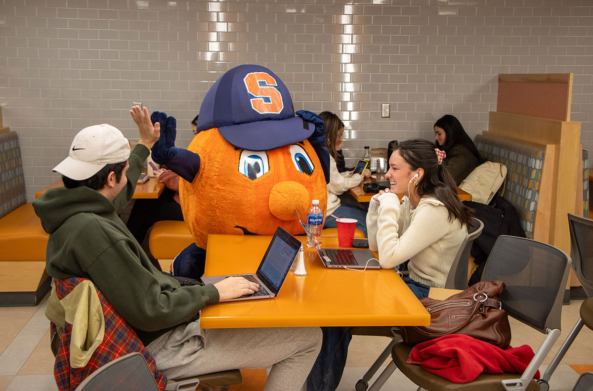 Otto the Orange standing next to a table with two people