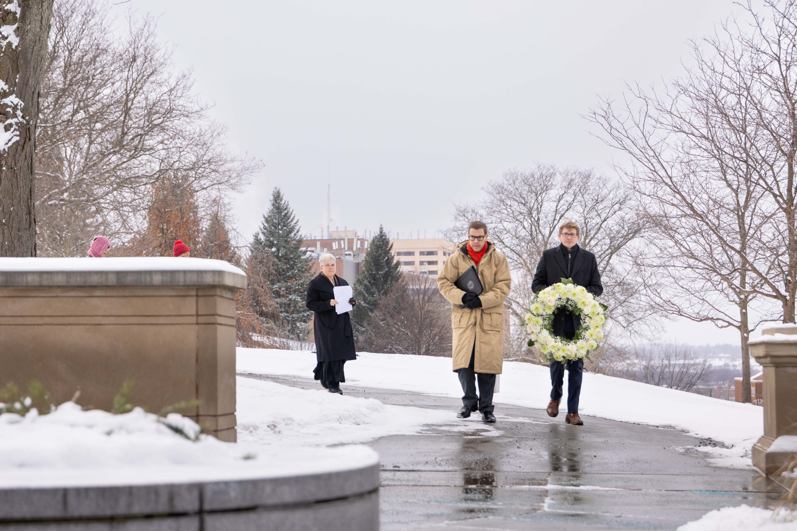 Three individuals walking on a snowy path, with one carrying a wreath. They are dressed in winter attire and appear to be in a solemn procession.