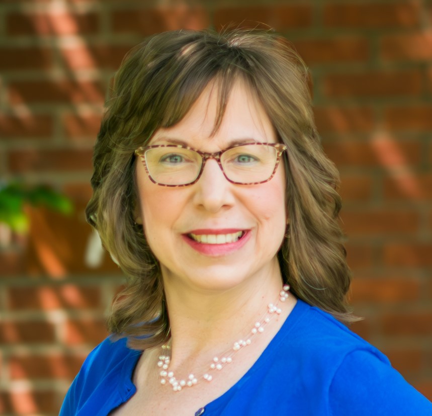 Person with wavy shoulder-length hair in blue top and pearl necklace standing in front of a brick wall.