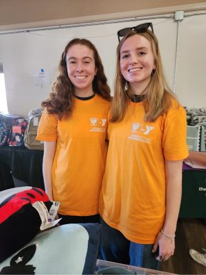 Two individuals are standing side by side, both wearing orange T-shirts with white logos and text on the front. The background shows a table with various items, including bags and folded clothes.