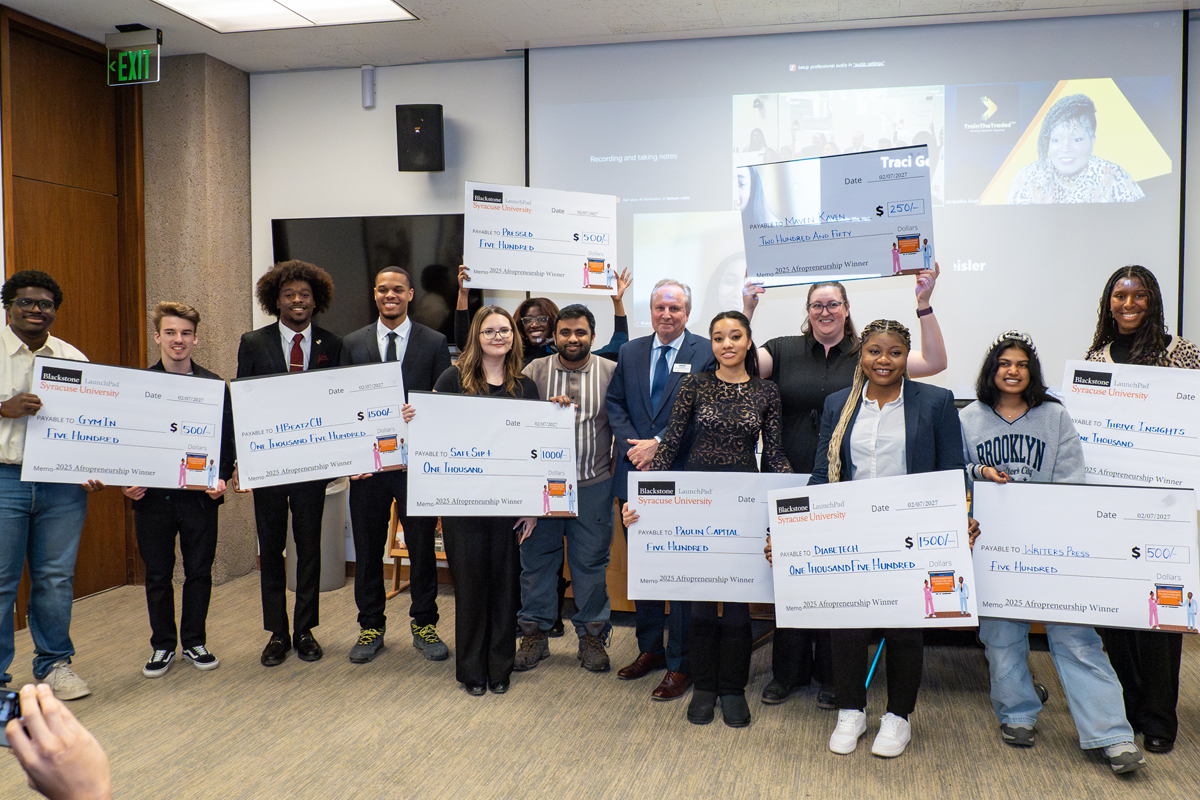 The image shows a group of eleven people standing in a room, each holding large ceremonial checks. The checks display various amounts of money, with most showing $1,000. The individuals are dressed in business casual attire. In the background, there is a screen displaying a video call or presentation. This appears to be an award or recognition event.