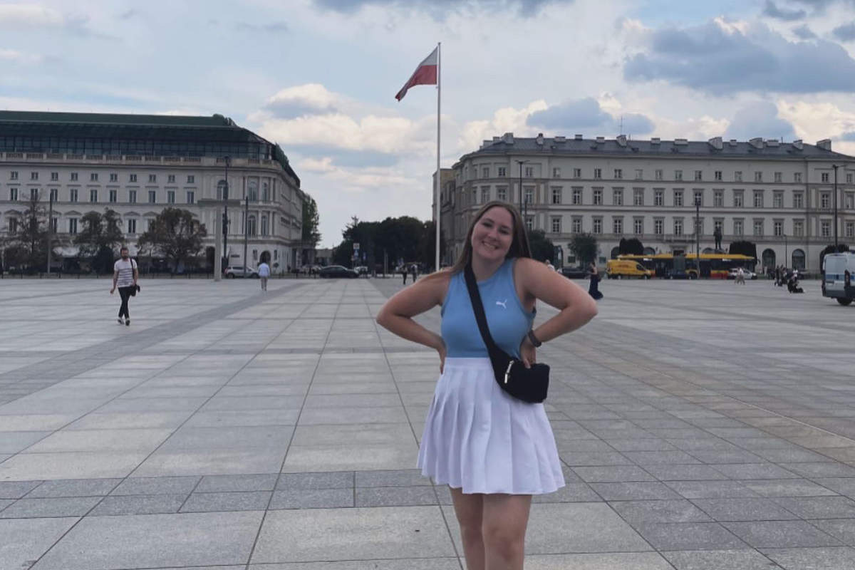 The image shows a person standing in a large, open square with tiled pavement. The person is wearing a blue top and a white skirt, with their hands on their hips and facing away from the camera. In the background, there are two large buildings with classical architecture, separated by a row of trees. A flagpole with a red and white flag stands prominently between the buildings. The sky above is partly cloudy.