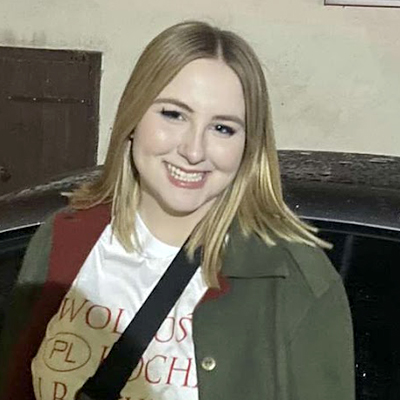 The image shows a person with shoulder-length blonde hair standing in front of a dark-colored car. They are wearing a white shirt with red text partially visible, an olive green jacket, and a black strap across their chest. In the background, there is a brown door and a light-colored wall.