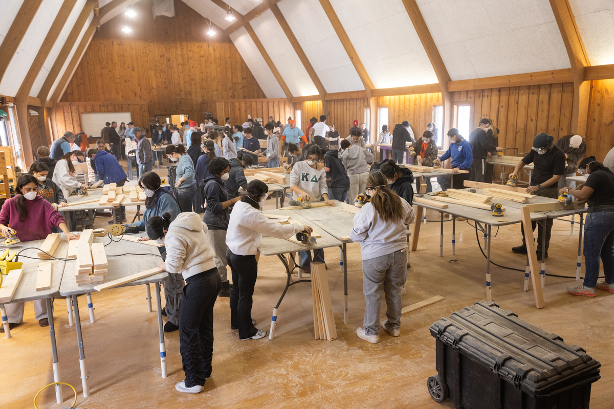 The image shows a large group of people working in a spacious, well-lit workshop with high ceilings and wooden walls. The individuals are engaged in various woodworking activities at multiple tables, using tools such as saws and sanders. The room is filled with workbenches, materials, and equipment. Everyone appears focused on their tasks, creating an atmosphere of industrious activity.