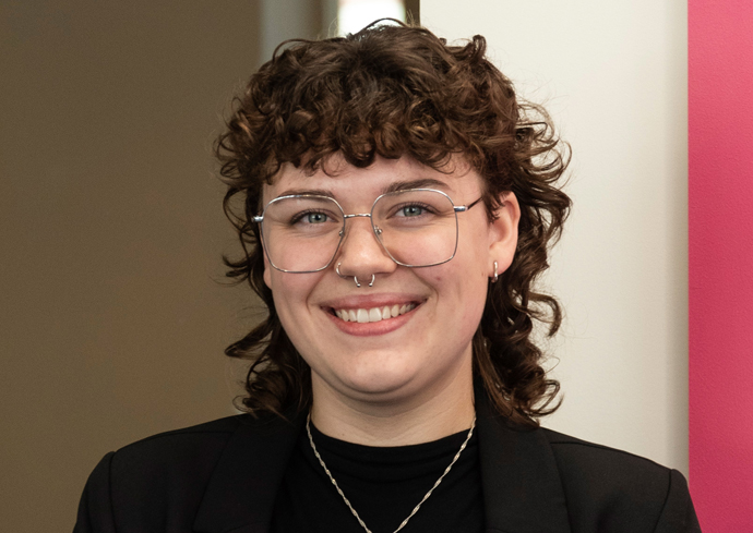 The image shows a person with curly hair wearing a black blazer over a black top and a silver necklace. The background consists of a beige wall on the left and a pink wall on the right.