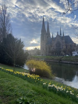 A church in Strasbourg, France, with a river running in front and clouds in the background.