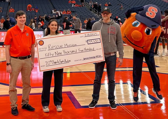 A group of four individuals is standing on a basketball court. Three people are holding a large ceremonial check made out to "Rescue Mission" for the amount of $59,500, dated 2/18/2023. The check is labeled "20th Charity Sports Auction." The fourth individual is dressed in an orange mascot costume with a blue hat and is posing playfully next to the group.