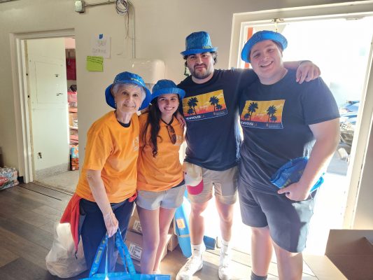 Four people standing together in a room, all wearing blue hats and holding blue bags. Two of them are in orange shirts, while the other two are in dark blue shirts with a palm tree design on the front. The room has wooden flooring, and an open door reveals another room with shelves containing various items. A green sign is visible on the wall near the door.