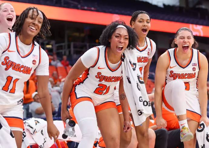 our basketball players in white and orange uniforms with "Syracuse" written on the front are seen in a dynamic pose, possibly during a game or practice. They are holding towels and appear to be focused and ready for action. The background shows an indoor stadium with spectators and a large screen displaying information.