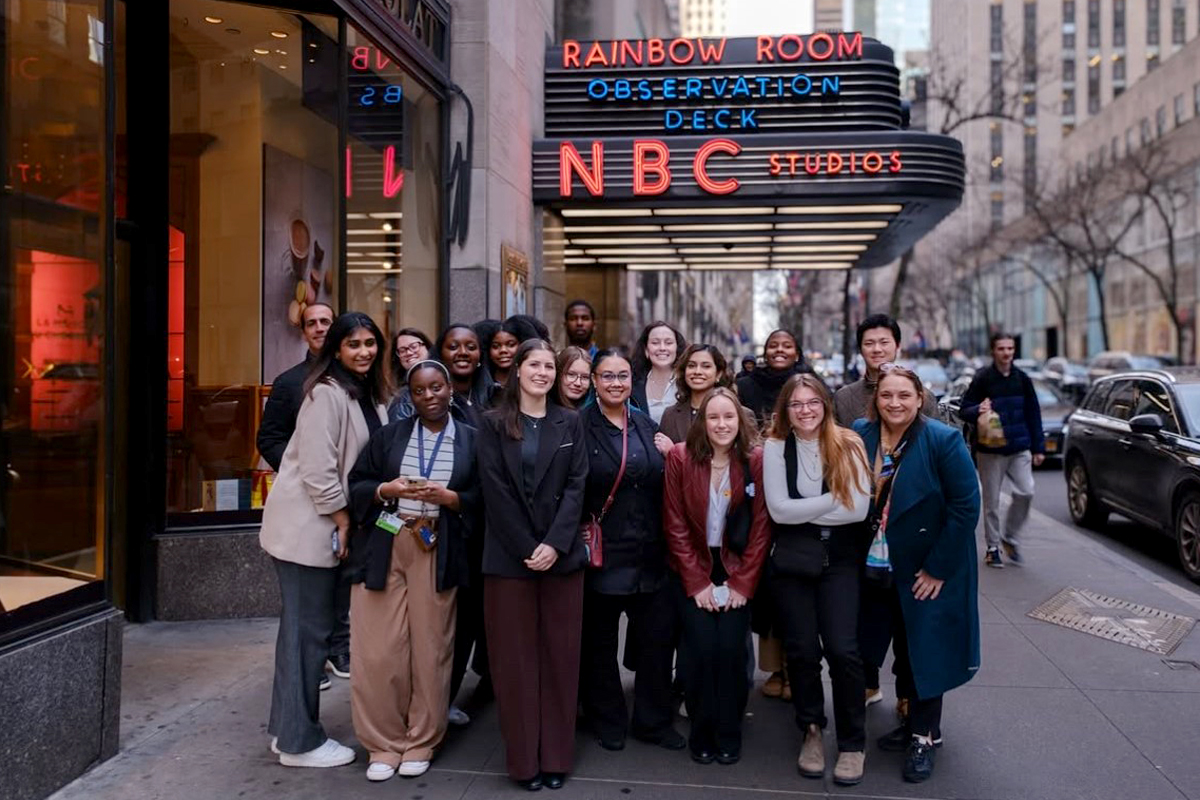 The image shows a group of people standing in front of the NBC Studios entrance at Rockefeller Center in New York City. The marquee above them reads "Rainbow Room Observation Deck NBC Studios." The group appears to be posing for a photo on the sidewalk, with some people wearing lanyards around their necks. The background shows a busy street with other pedestrians and tall buildings.