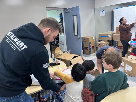 A group of people is in a room filled with cardboard boxes. One person, wearing a black hoodie with the text 'STACKMART TEAM LEAD' on the back, is handing an item to a child. Another adult and two children are also visible, engaged in sorting or packing items. In the background, there is a whiteboard and more boxes stacked against the walls. The scene appears to be an organized activity involving packing or distributing goods.