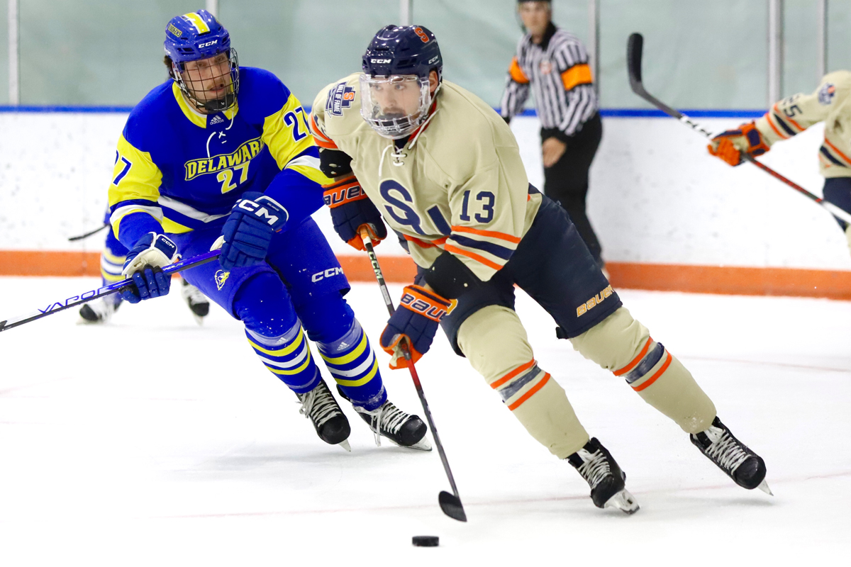 The image shows two ice hockey players in action on the rink. The player on the left is wearing a blue and yellow uniform with "DELAWARE" written across the chest and the number 27 on the jersey. The player on the right is wearing a beige uniform with blue and orange stripes, marked with the number 13. Both players are focused on controlling or chasing after a black hockey puck. An official in black and white stripes is visible in the background, watching the play closely.