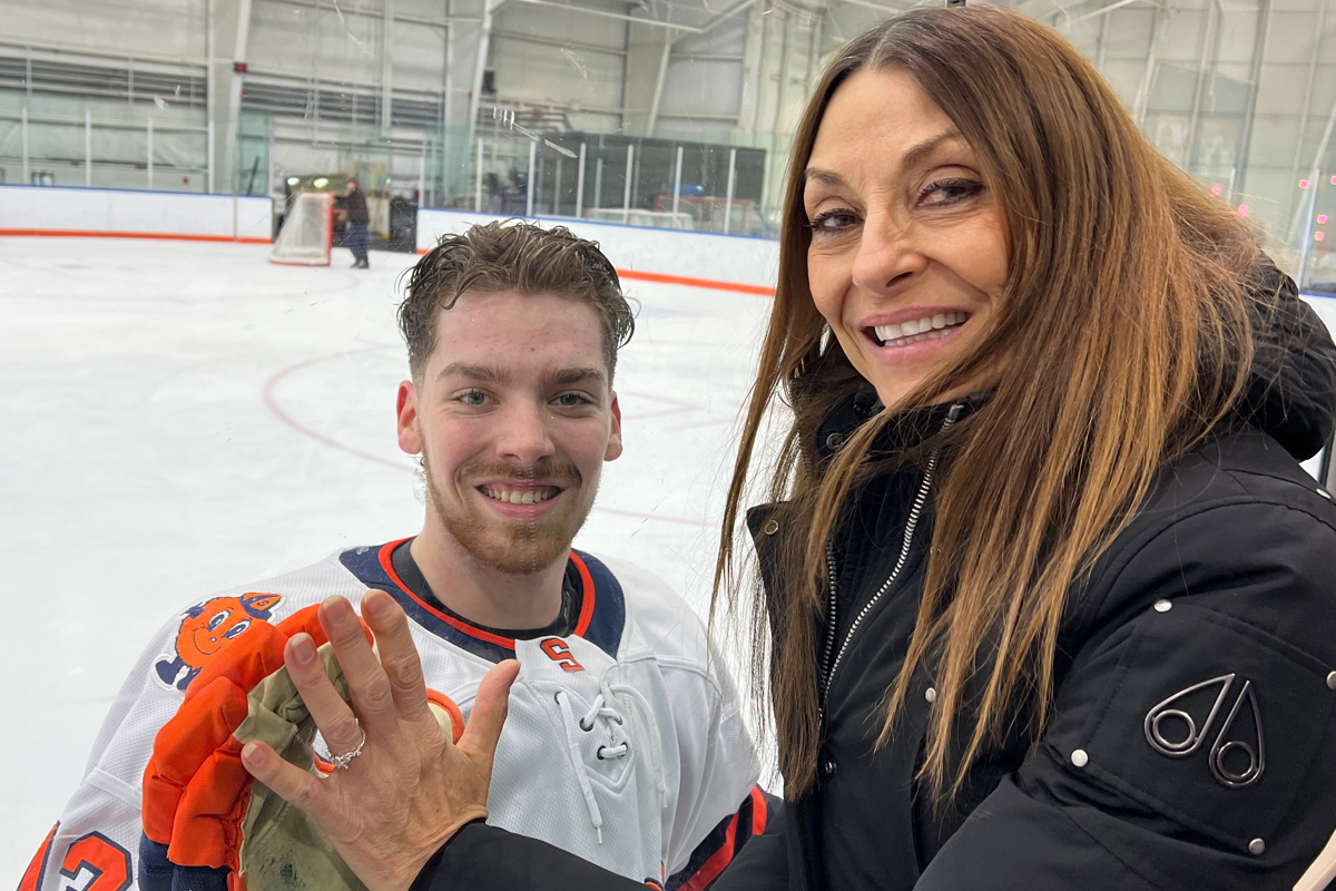 The image shows a hockey player in a white jersey with orange and blue accents standing on an ice rink. The player is holding a hockey glove. Next to the player, there is another person wearing a black jacket. The background shows the ice rink, boards, and part of the goal net.