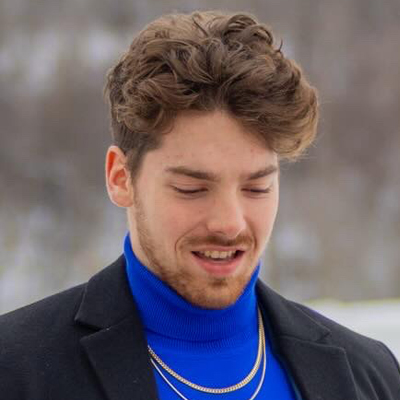 The image shows a person with curly brown hair wearing a blue turtleneck sweater, a black blazer, and two gold chains around the neck. The background appears to be an outdoor setting with blurred trees.