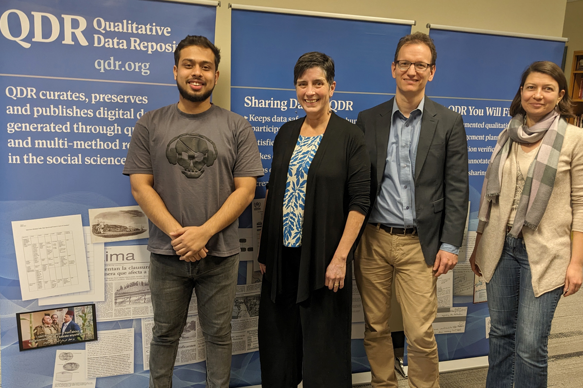 The image shows four individuals standing in front of two blue banners with text and images related to the Qualitative Data Repository (QDR). The banners contain information about QDR's mission, which includes curating, preserving, and publishing digital data generated through qualitative and multi-method research in the social sciences. The individuals are dressed in casual to business-casual attire.