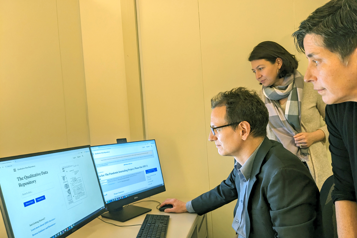 The image shows three people in an office setting, looking at two computer monitors on a desk. The person seated is using a mouse and keyboard, while the other two stand behind. The left monitor displays "The Qualitative Data Repository" with some text and images, while the right monitor shows a webpage titled "How to Use the Qualitative Data Repository (QDR) for Research."