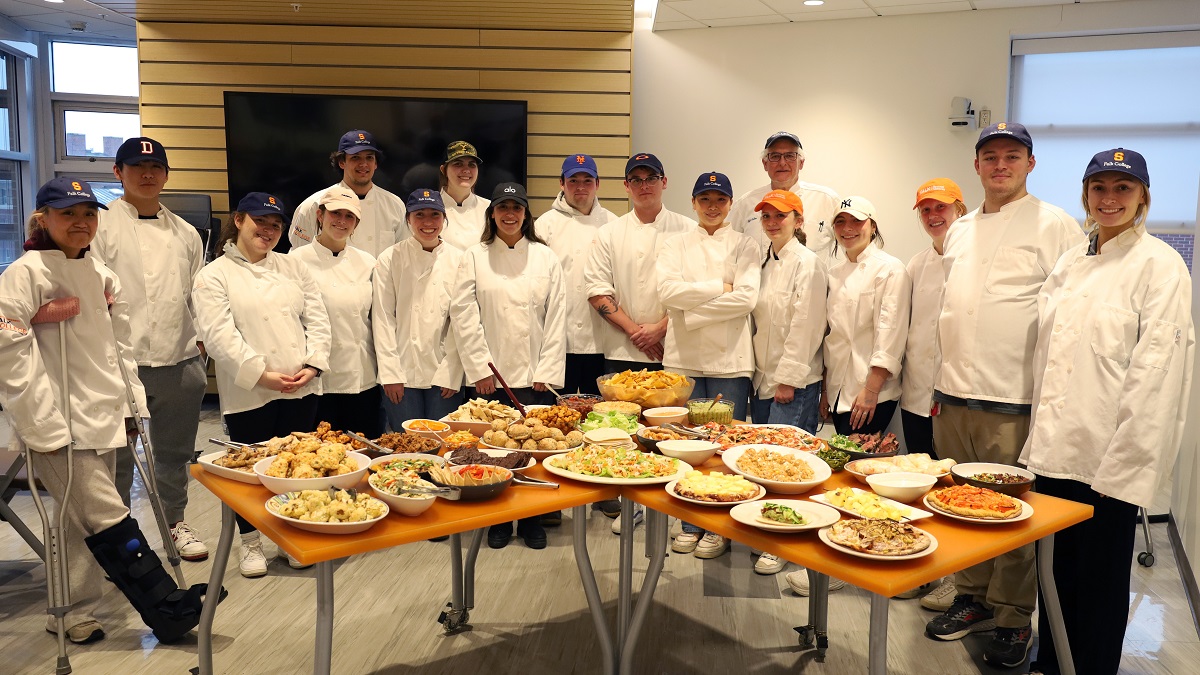 group of students in chef's attire standing behind a table filled with food items