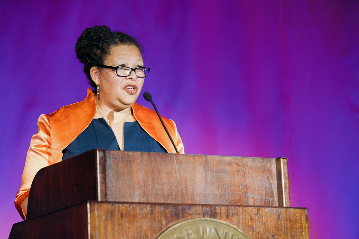 The image shows a person standing behind a wooden podium, speaking into a microphone. The individual is dressed in an orange and blue outfit, and the backdrop is bright purple.