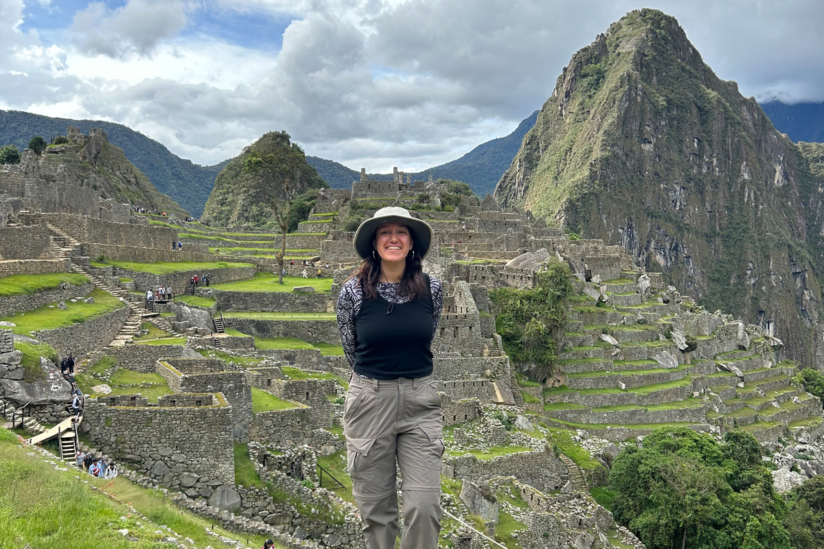 The image shows a person standing in front of the ancient Incan city of Machu Picchu, located high in the Andes Mountains of Peru. The background features terraced fields, stone structures, and steep mountain peaks under a partly cloudy sky. The person is wearing a black hat and light-colored clothing.