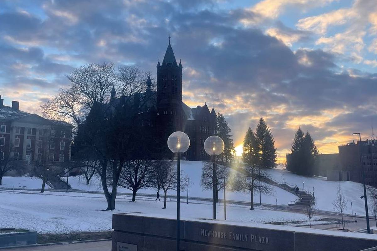 The image shows a scenic view of a snow-covered campus at sunrise or sunset. The sky is partly cloudy with hues of blue, orange, and yellow. In the background, there is a large, historic building with a tall spire and multiple turrets silhouetted against the sky. Bare trees are scattered around the snowy landscape. In the foreground, there are two spherical street lamps on poles and a stone sign that reads "Newhouse Family Plaza."