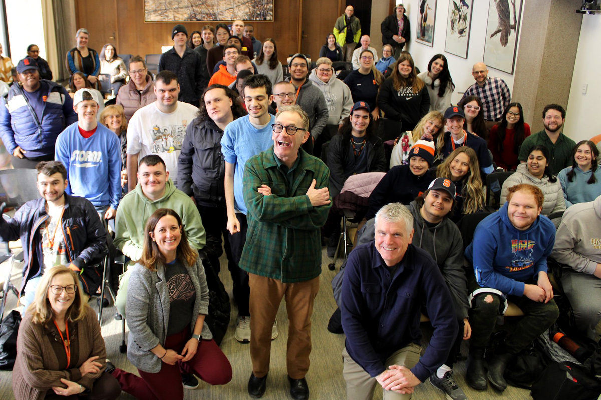 The image shows a large group of people gathered in a room with wooden walls and framed pictures on one side. Some individuals are seated while others stand, facing the camera. A few people in the back are wearing high-visibility vests.