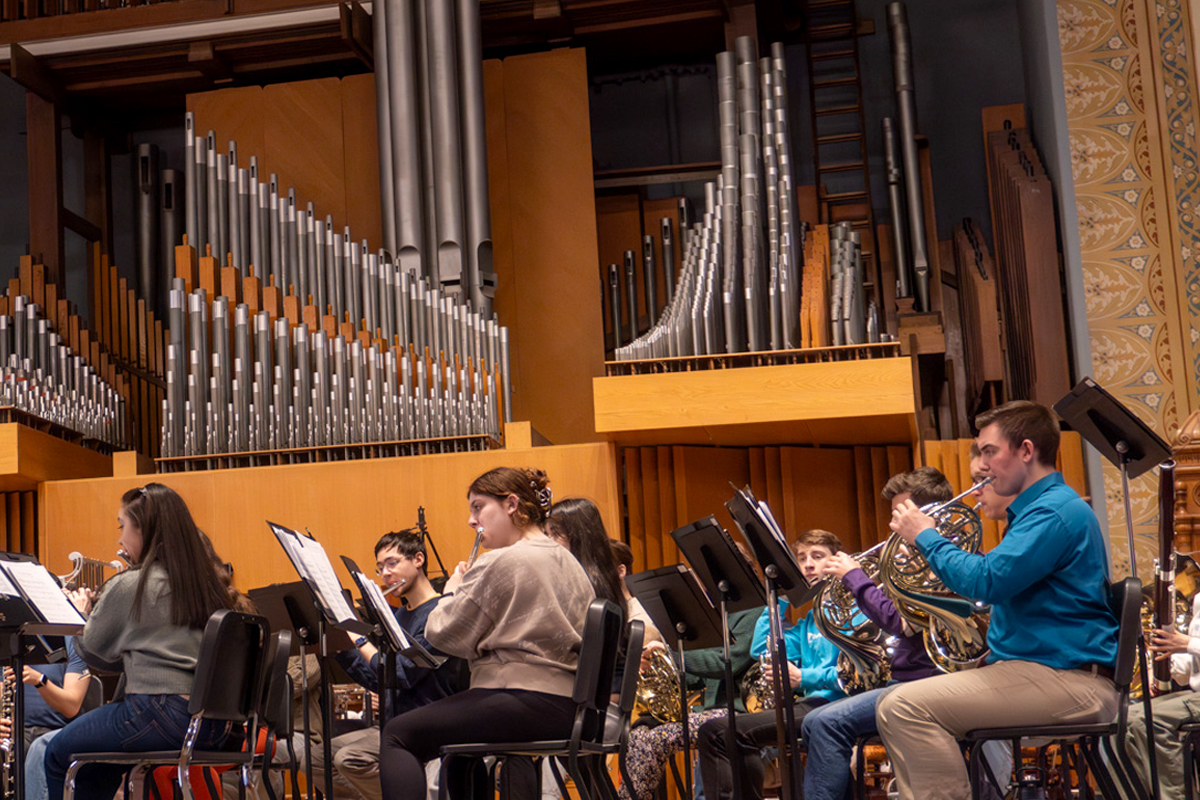 The image shows a group of musicians seated in a rehearsal space, playing various brass and woodwind instruments. They are arranged in rows, facing music stands with sheet music. Behind them is a large pipe organ with numerous pipes of varying lengths. The setting appears to be an auditorium or concert hall with wooden paneling and decorative elements on the walls.