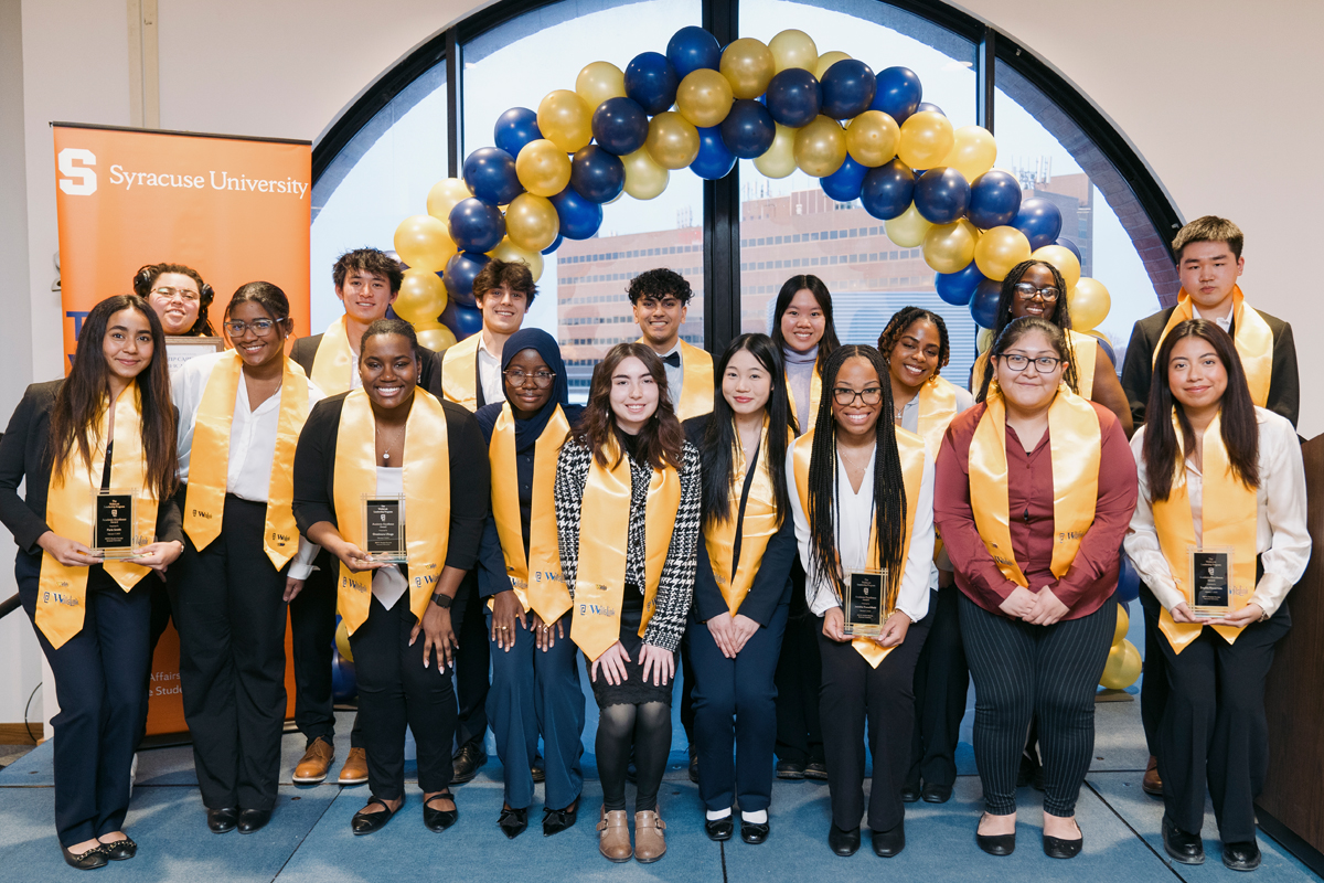 A group of people are standing and kneeling in two rows, posing for a photo in front of an arch made of blue and yellow balloons. They are wearing formal attire with yellow stoles around their necks. Behind them is a large window showing a building outside. To the left, there is an orange banner with "Syracuse University" written on it.