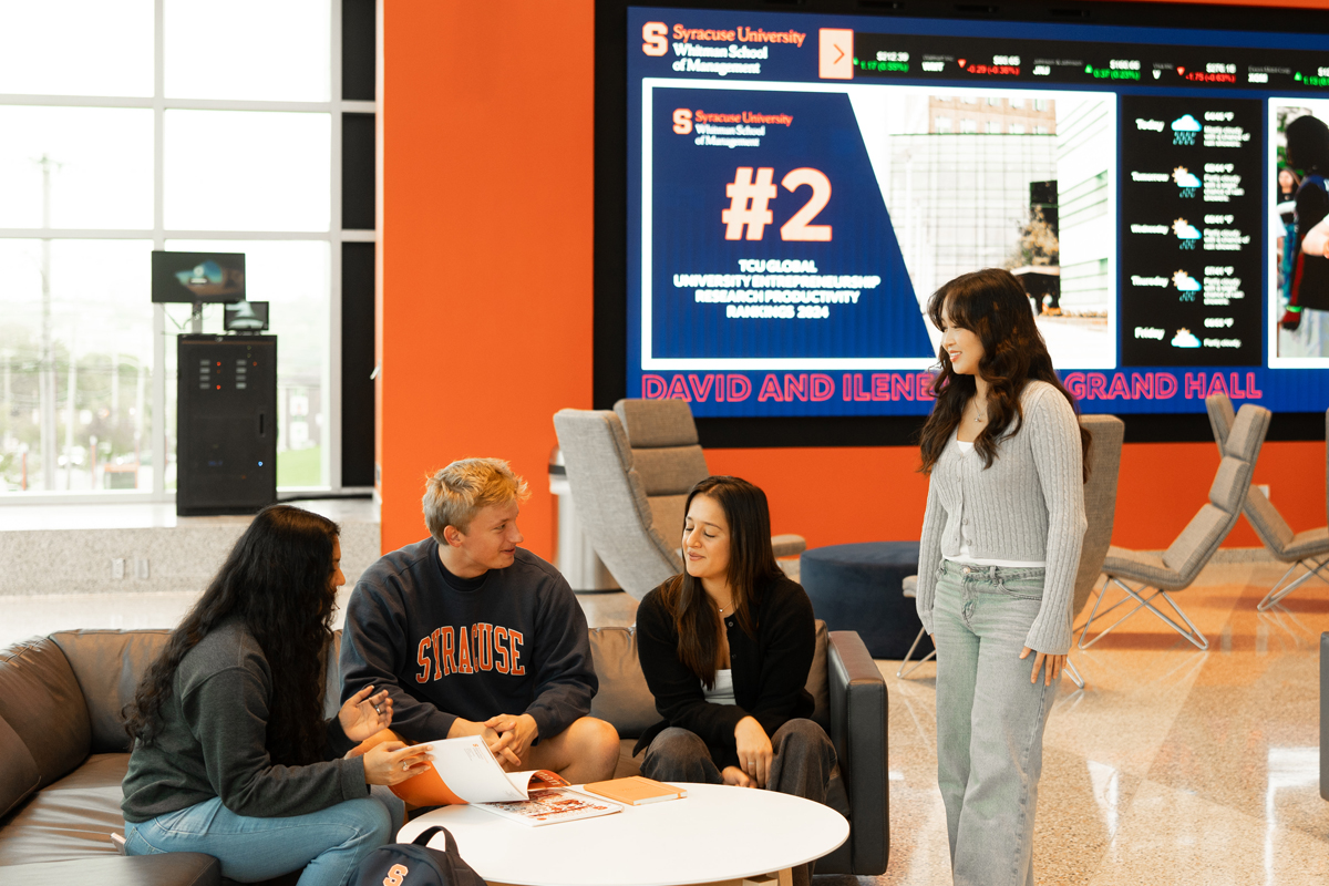 The image shows four students interacting in a lounge area with an orange wall and a large digital display screen behind them. The display features Syracuse University information, highlighting its #2 ranking for top global universities for industrial engineering and management. The students are seated on gray couches around a white circular table, discussing something while looking at papers.