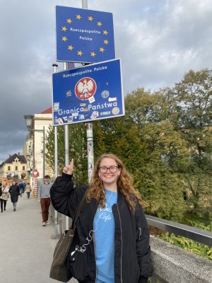 The image shows a person standing on a bridge, pointing at a blue sign with yellow stars and white text that reads "Rzeczpospolita Polska." Below this sign, there is another blue sign with the same text and an emblem of a white eagle. The lower sign also includes the words "Granica Państwa" and various stickers. The background shows trees, buildings, and several other people walking.