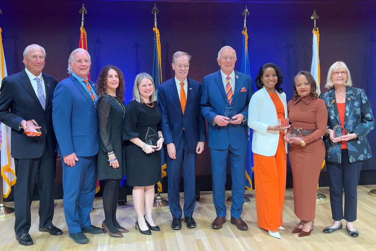 The image shows a group of eight people standing in a row on a stage, holding awards. The background features several flags and a dark backdrop with blue lighting. The individuals are dressed formally, with men in suits and women in dresses or business attire.