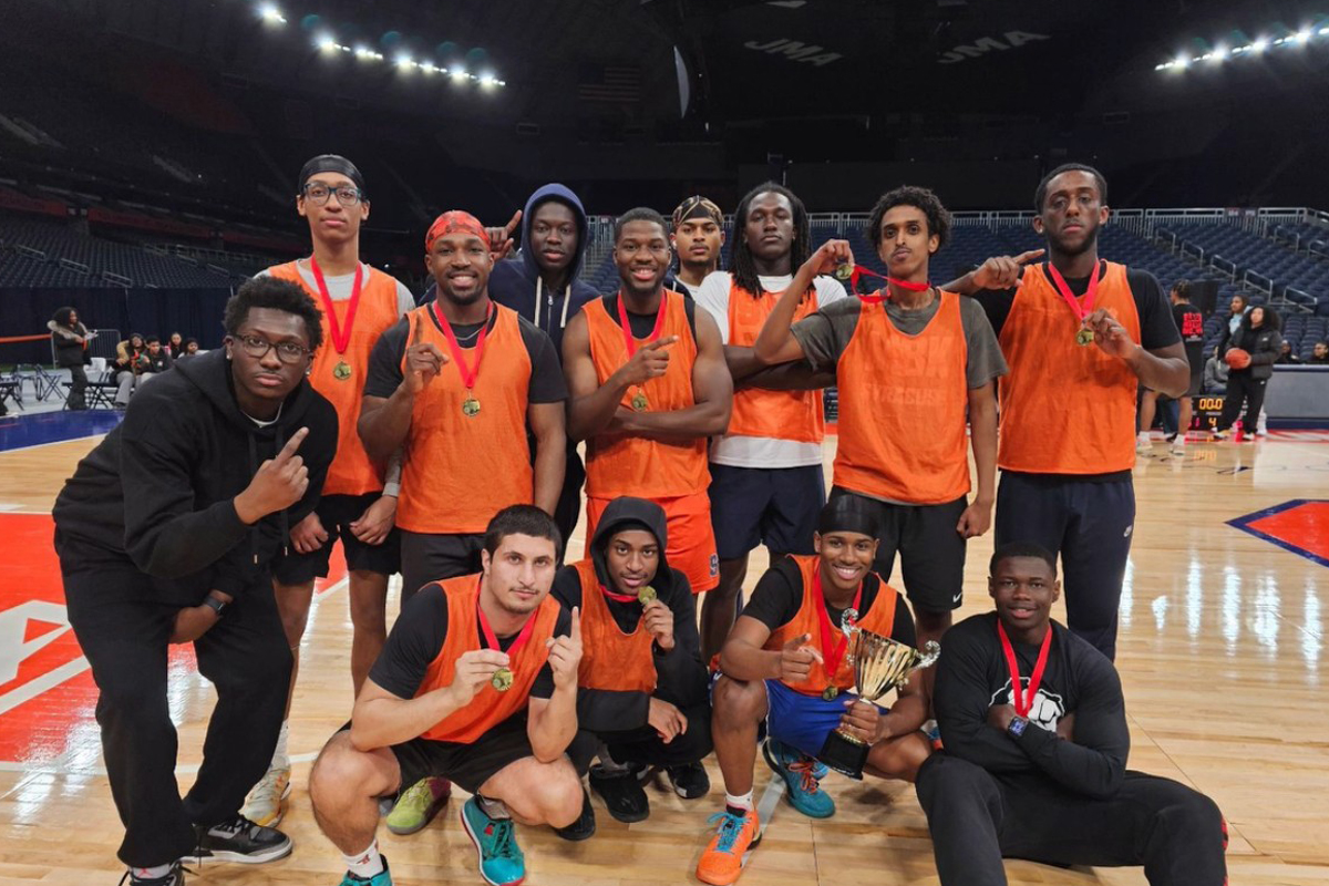 The image shows a group of twelve people posing on an indoor basketball court. They are wearing orange bibs and holding medals, with one person in the front holding a trophy. The background shows empty stadium seats and a large scoreboard.